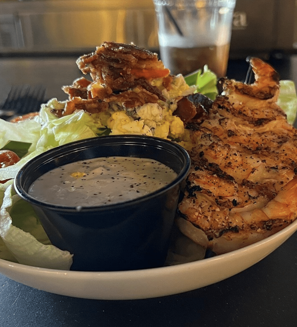 A shrimp-topped salad with chopped bacon, eggs, lettuce, tomatoes, and a cup of dressing.